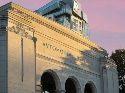 Close-up exterior view of the Automotive Building at Exhibition Place in Toronto during sunset. The historic stone façade features three large archways, decorative Art Deco-inspired detailing, and the words “Automotive Building” engraved above the entrance.