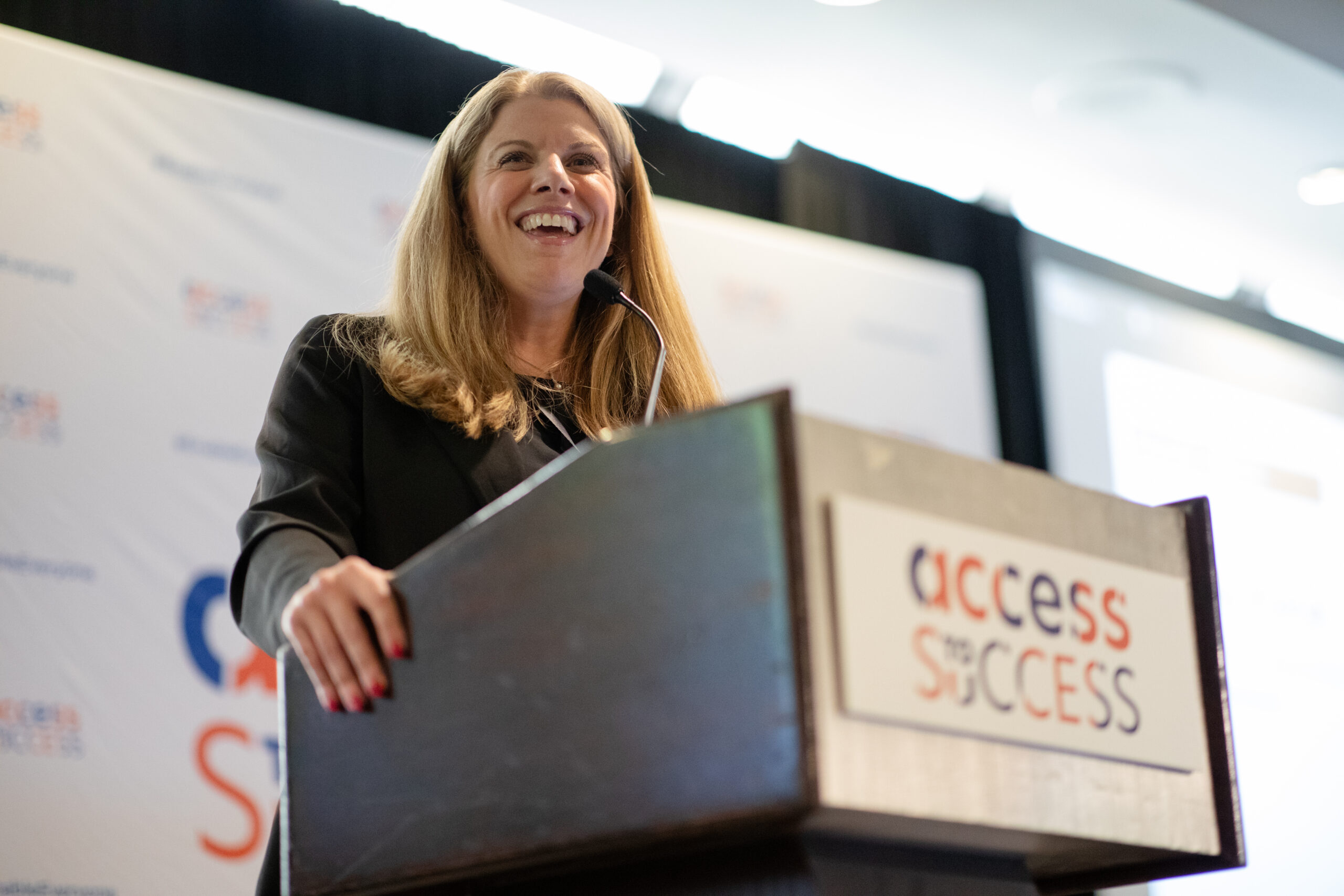 Regina “Gina” Kline speaks at an Access to Success (ATS) event, standing behind a podium branded with the ATS logo. She addresses the audience with a microphone, smiling as she delivers remarks. An ATS event backdrop is visible behind her, reinforcing the focus on accessibility innovation, entrepreneurship, and ecosystem collaboration.