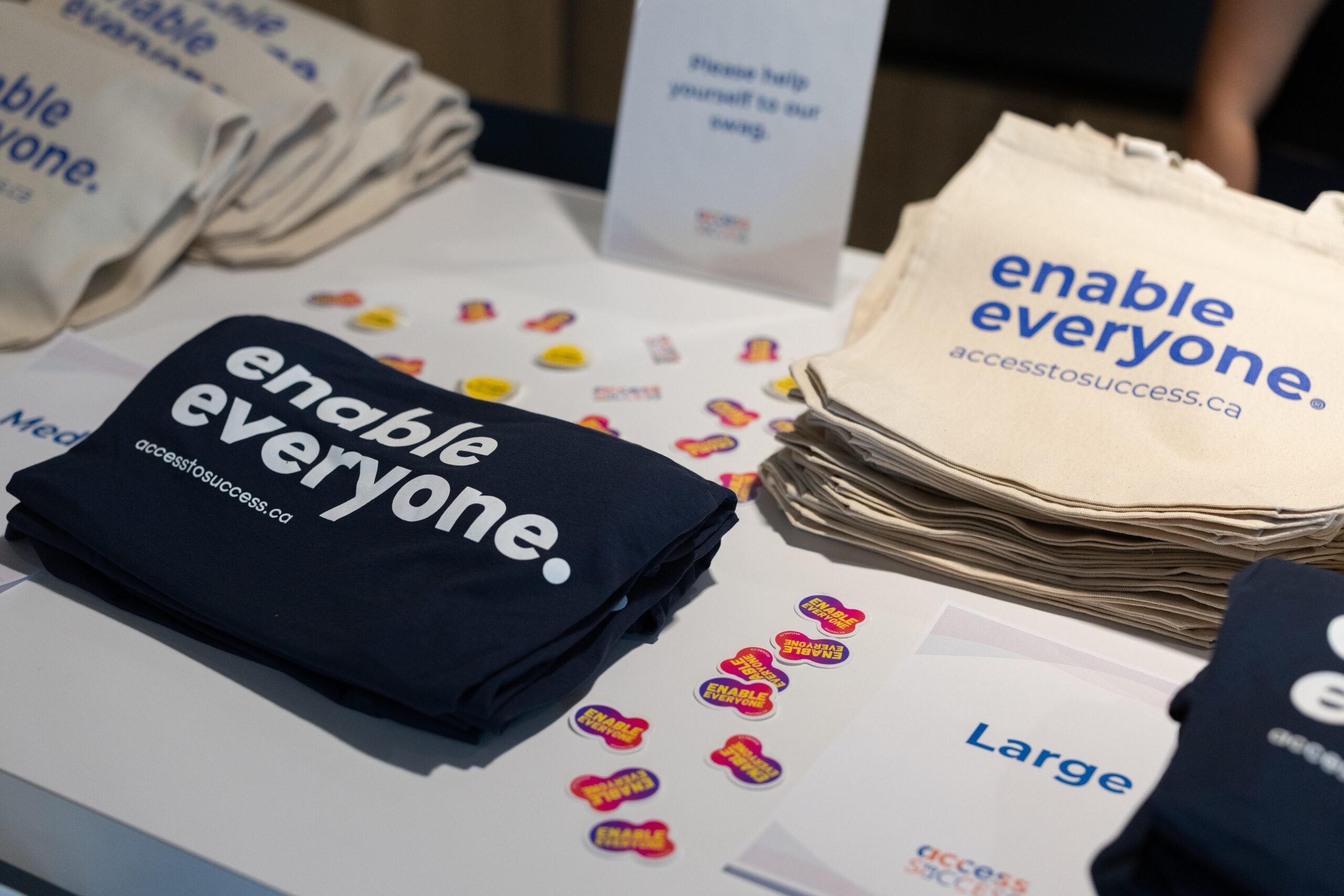 Table display featuring Access to Success (ATS) promotional materials. Stacks of navy and natural-coloured tote bags printed with the “Enable Everyone” message and the ATS website are arranged on a table alongside branded stickers. A sign invites attendees to help themselves to the items, with additional size labels visible for event merchandise.