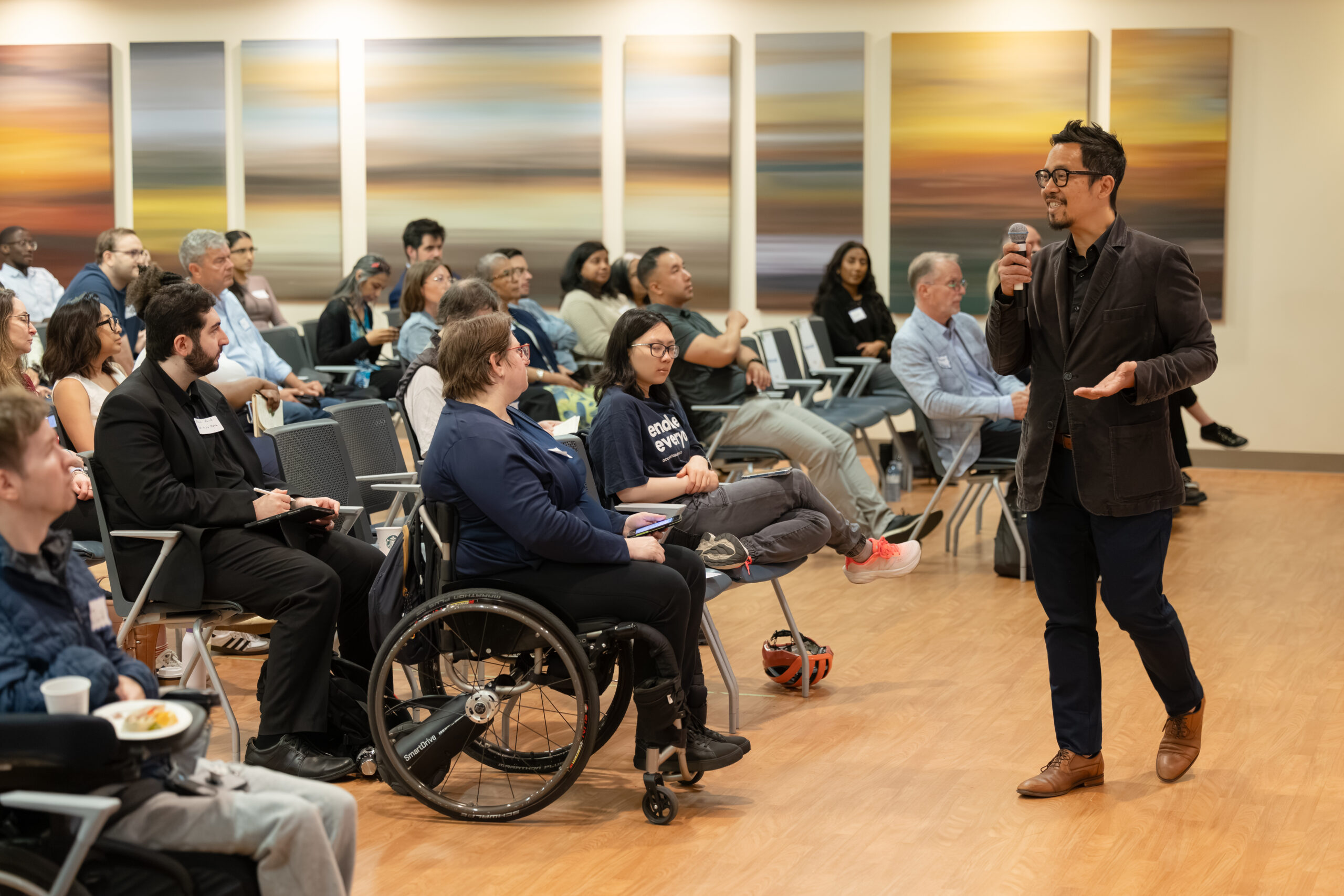 Anthony Palma speaks to attendees during AIX 2026, holding a handheld microphone and gesturing as he addresses the audience. Attendees, including a wheelchair user seated in the front row, listen attentively in a bright conference room with abstract wall art.