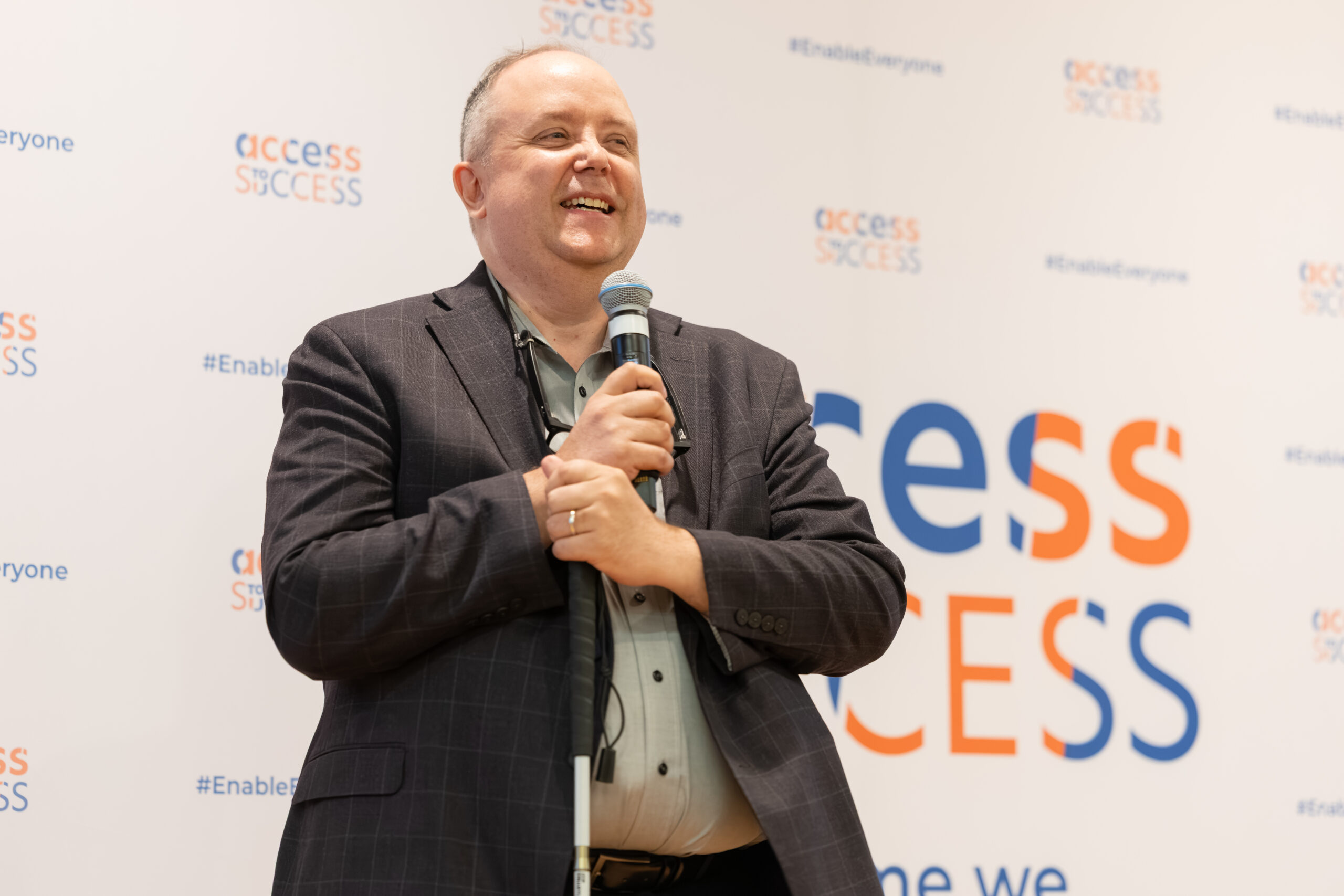 Wayne Henshall of CNIB Foundation speaks at AIX 2026. Standing in front of an Access to Success (ATS)-branded backdrop, he smiles while holding a microphone and a white cane during his presentation on accessibility innovation.