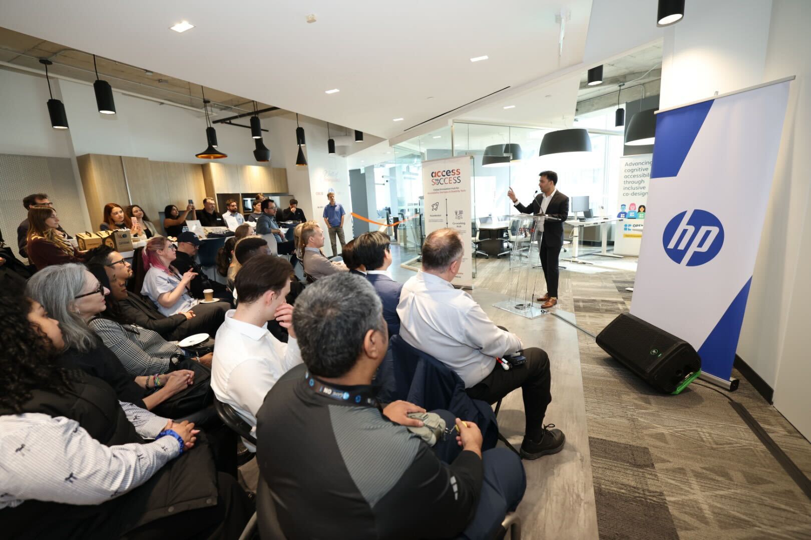 Varun Chandak speaks to an audience at an event hosted at the ATS Innovation Hub. Standing at a podium, he presents to attendees gathered in the coworking space, with ATS and HP banners displayed beside him. Audience members are seated throughout the room, listening to the presentation, while glass-walled meeting rooms and collaborative workspaces are visible in the background.