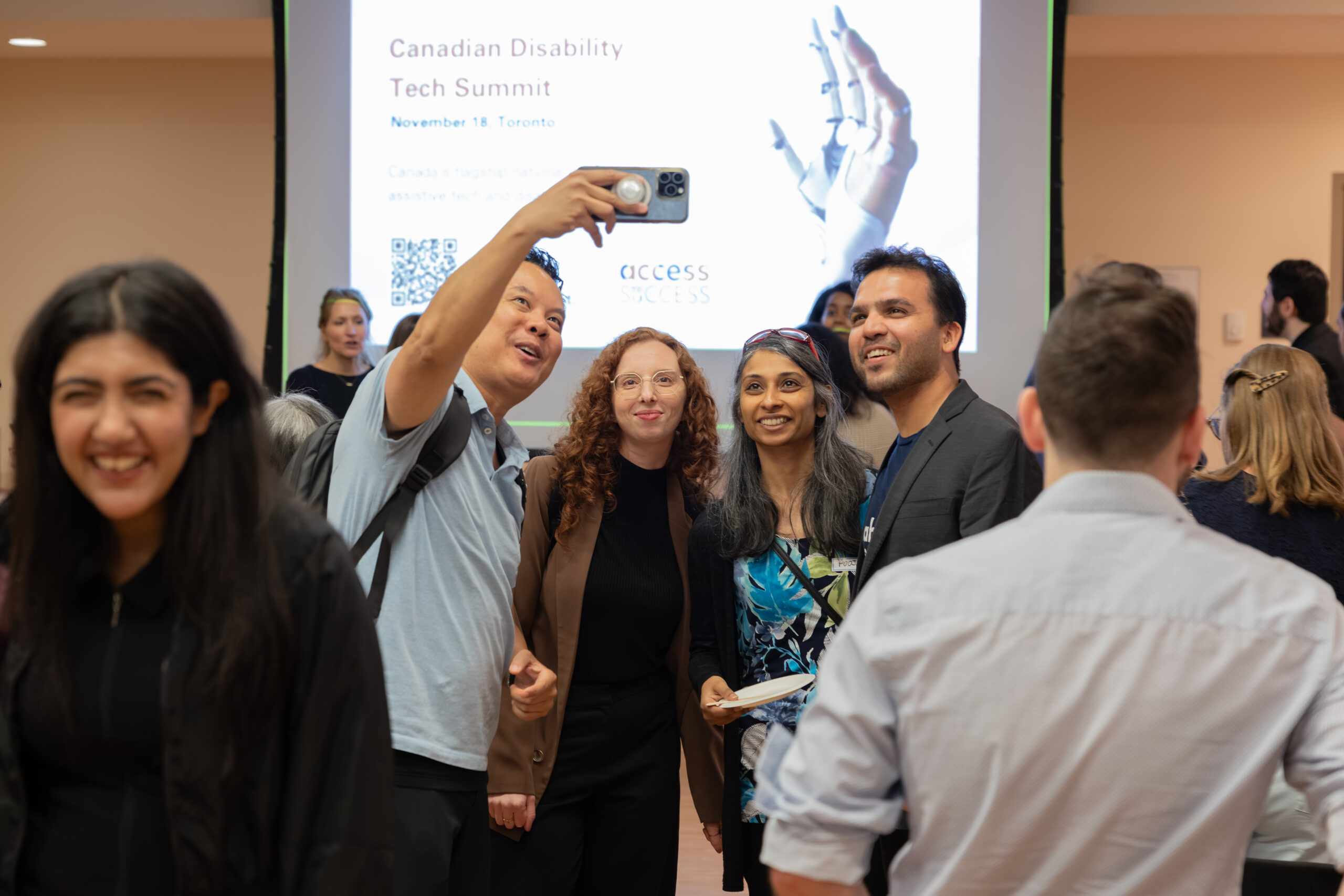 Attendees gather and chat during an Access to Success event. In the centre, four people smile while one holds up a smartphone to take a group selfie. A large screen behind them displays the Canadian Disability Tech Summit, and other attendees mingle throughout the room.