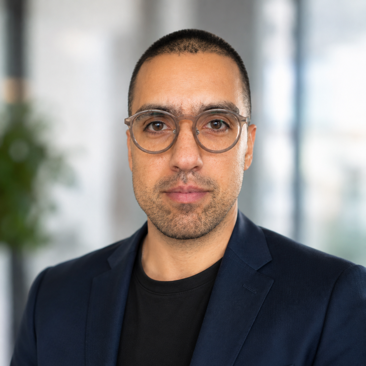 Professional headshot of Omid Sadeghi wearing a navy blazer, black shirt, and round glasses, facing the camera against a blurred office background.