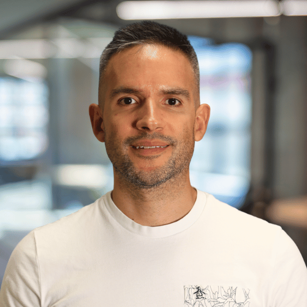 Professional headshot of Juan Pablo Carstens wearing a white T-shirt, smiling at the camera against a blurred office background.