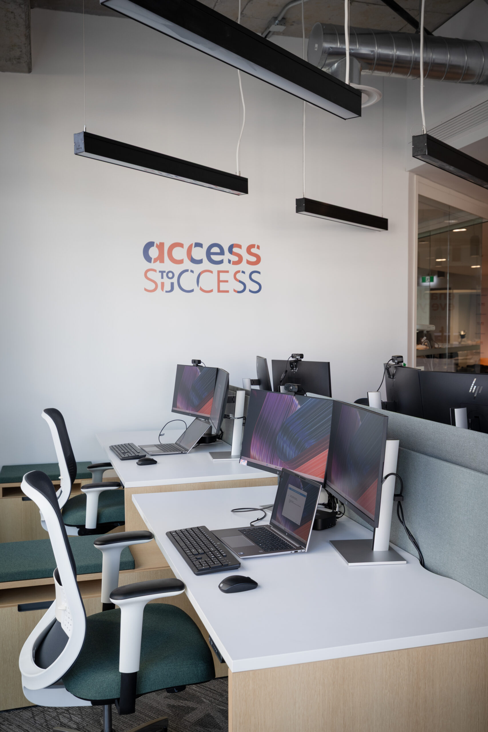 A workspace inside the Access to Success Innovation Hub features shared desks with laptops, dual monitors, keyboards, ergonomic chairs, and webcams for virtual collaboration. The Access to Success logo is displayed on the wall